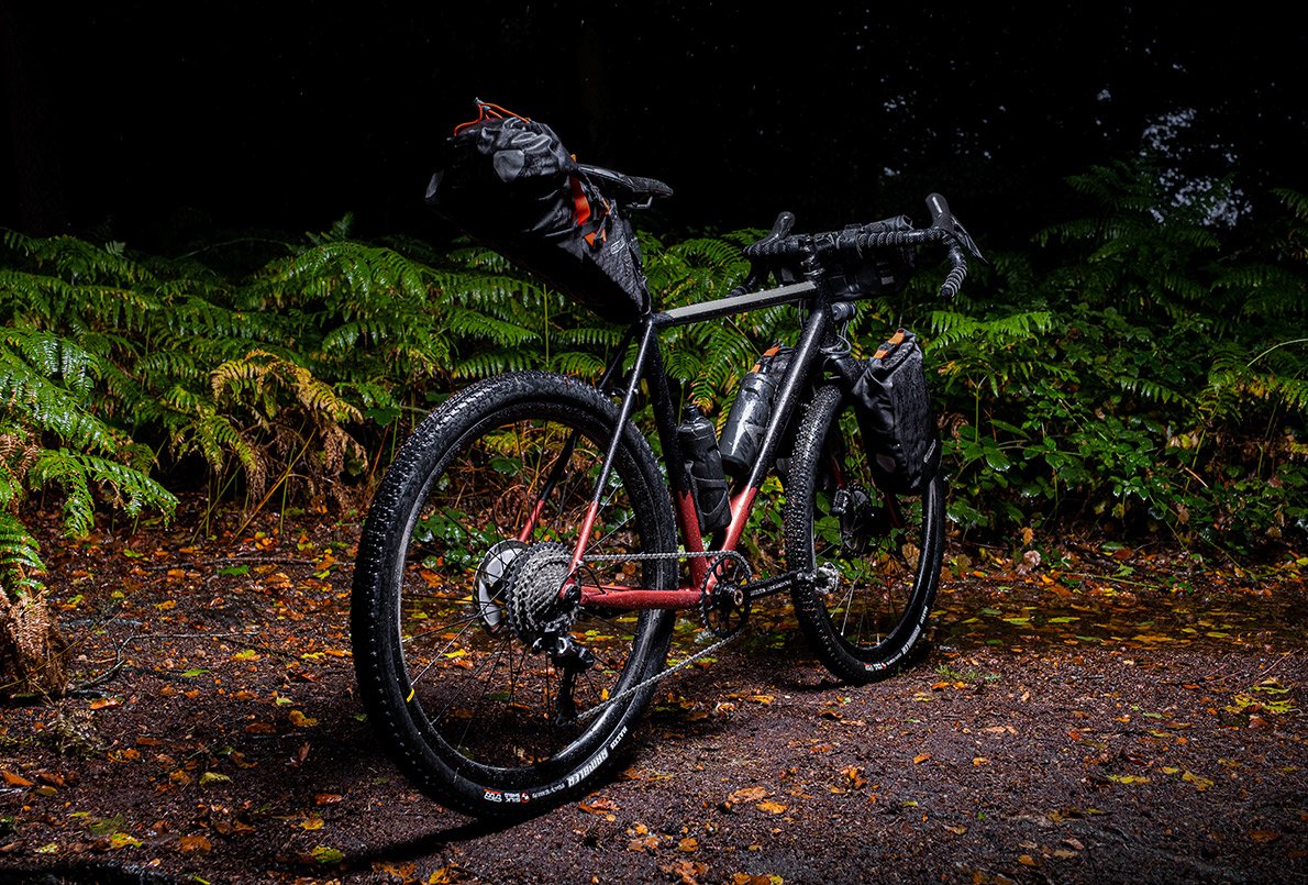 Vélo Wish One Quest Gravel, noir et cuivre, face arrière, avec équipement bikepacking Ortlieb sur un chemin en forêt