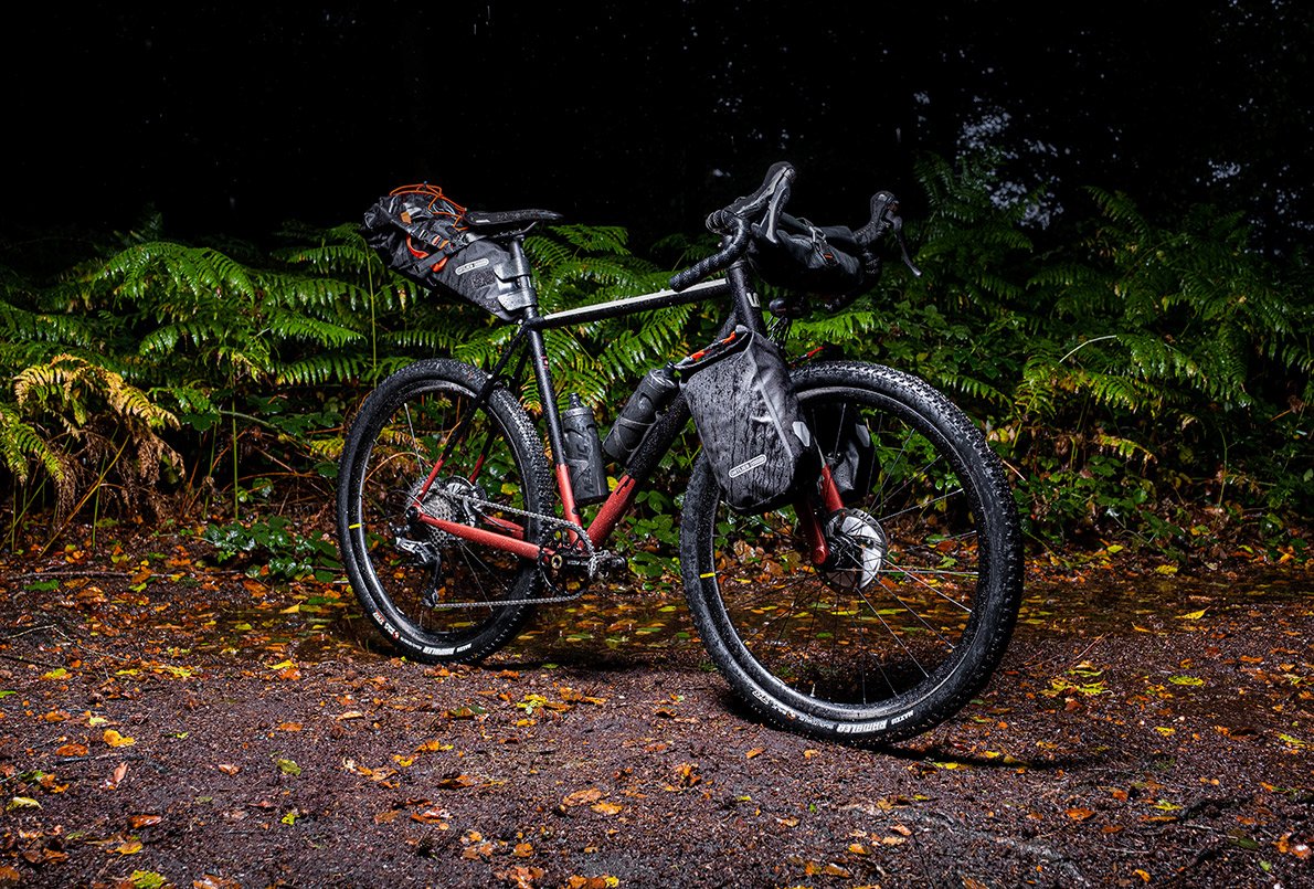 Vélo Wish One Quest Gravel noir et cuivre, en entier, avec équipement bikepacking Ortlieb sur un chemin en forêt
