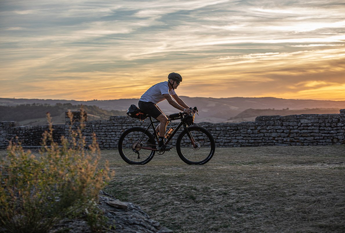 Cycliste sur le vélo gravel Wish One Quest équipé bikepacking roulant sur chemin avec des collines en arrière-plan