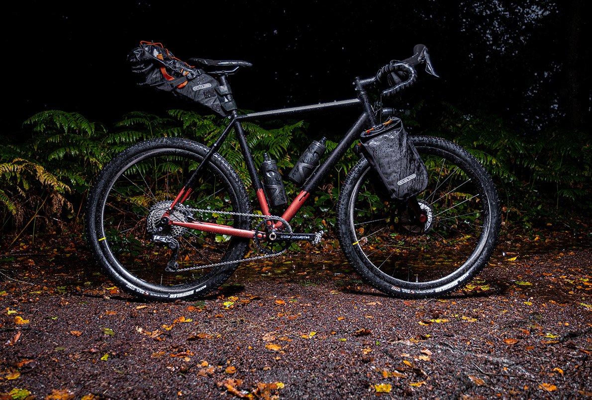 Vélo Wish One Quest Gravel noir et cuivre, de profil, avec équipement bikepacking Ortlieb sur un chemin en forêt