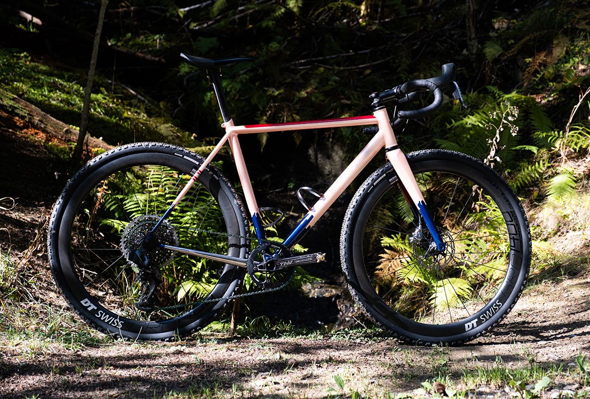 Vélo Wish One SUB Gravel rose, bleu marine et rouge, de profil sur un chemin en forêt