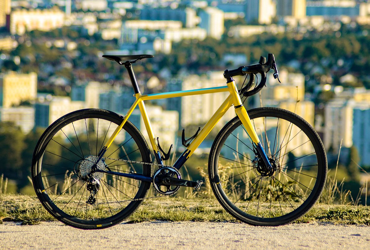 Vélo Wish One SUB Gravel jaune, bleu marine et vert de profil sur chemin avec la ville en arrière plan